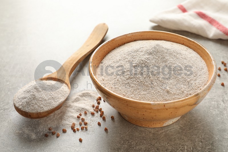Bowl and spoon of buckwheat flour with grains on grey table, space for text