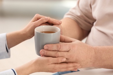 Nurse giving cup of tea to elderly man against blurred background, closeup. Assisting senior generation