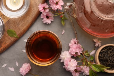 Photo of Traditional ceremony. Cup of brewed tea, teapot and sakura flowers on grey table, flat lay
