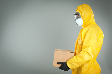 Man wearing chemical protective suit with cardboard box on light grey background, space for text. Prevention of virus spread