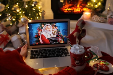 MYKOLAIV, UKRAINE - DECEMBER 23, 2020: Woman with sweet drink watching The Christmas Chronicles movie on laptop near fireplace at home, closeup. Cozy winter holidays atmosphere
