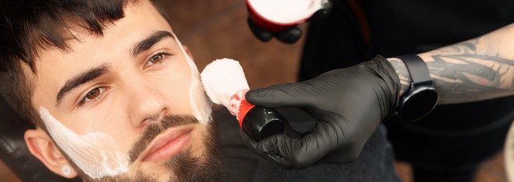 Image of Professional hairdresser applying shaving foam onto client's beard in barbershop, closeup. Banner design