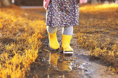 Photo of Little girl wearing rubber boots walking in puddle outdoors, closeup