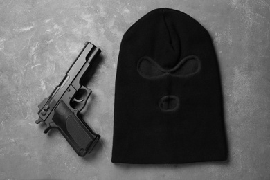 Black knitted balaclava and pistol on grey table, flat lay