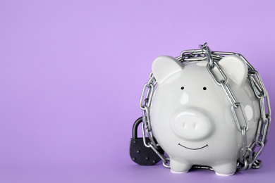 Piggy bank  with steel chain and padlock on lilac background, space for text. Money safety concept