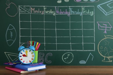 Alarm clock and stationery on wooden table near green chalkboard with drawn school timetable
