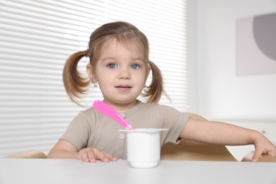 Photo of Cute little child eating tasty yogurt from plastic cup with spoon at white table indoors