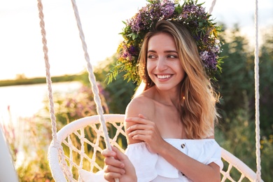 Young woman wearing wreath made of beautiful flowers on swing chair outdoors