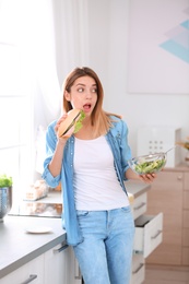 Photo of Beautiful young woman eating sandwich instead of salad in kitchen. Failed diet