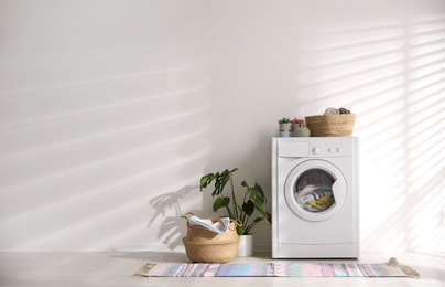 Modern washing machine and wicker basket with laundry near white wall, space for text. Interior design