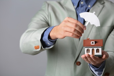 Male agent covering house model with umbrella cutout on grey background, closeup. Home insurance