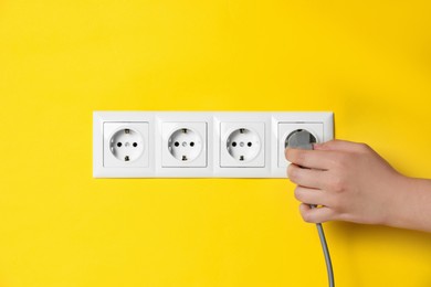 Woman inserting plug into power socket on yellow wall, closeup. Electrical supply