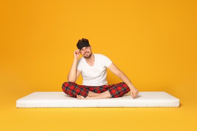 Smiling man in sleeping mask sitting on soft mattress against orange background