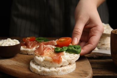 Woman holding puffed rice cake with prosciutto, tomato and basil at wooden table, closeup