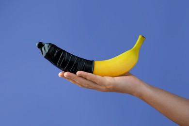 Woman holding banana in condom on light blue background, closeup. Safe sex concept