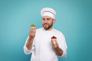 Photo of Happy professional confectioner in uniform holding delicious tartlets on light blue background