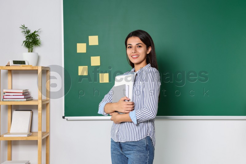 Happy young teacher with books at blackboard in classroom. Space for text
