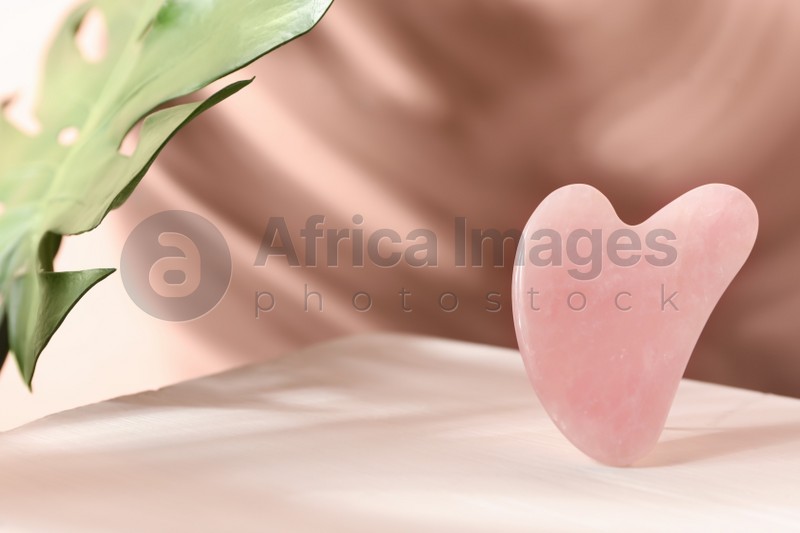 Photo of Rose quartz gua sha tool and tropical leaf on table, space for text