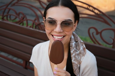 Beautiful young woman eating ice cream glazed in chocolate on bench outdoors