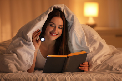 Young woman with flashlight reading book under blanket in bedroom