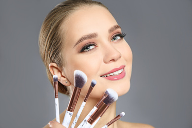 Beautiful woman with makeup brushes on light grey background