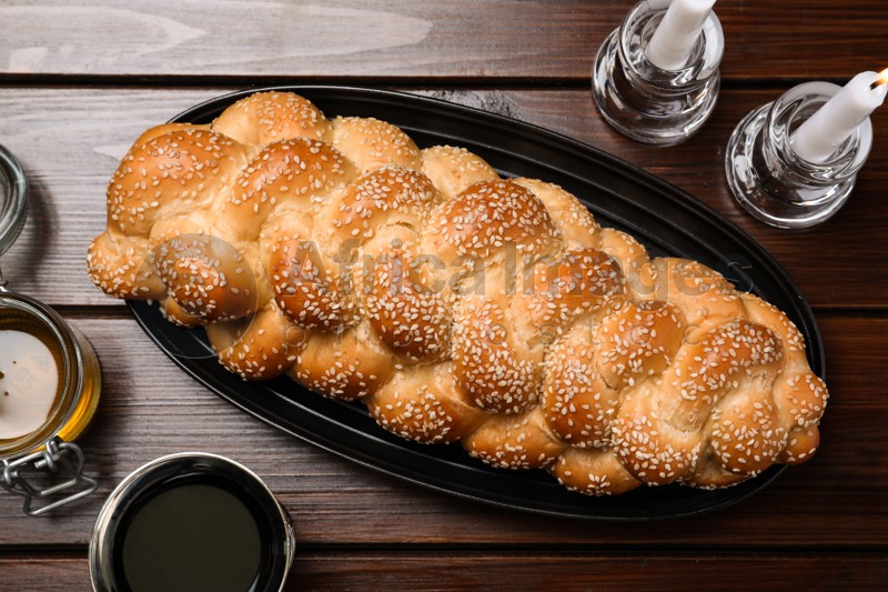 Homemade braided bread with sesame seeds, goblet, honey and candles on wooden table, flat lay. Traditional Shabbat challah