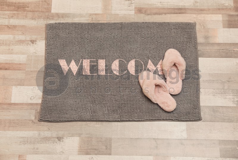 Door mat with word WELCOME and fluffy slippers on floor, top view