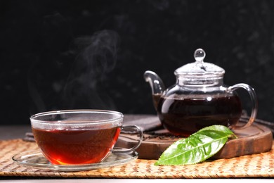 Photo of Aromatic hot tea in glass cup with saucer, teapot and leaves on table against black background. Space for text