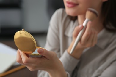 Photo of Young woman with cosmetic pocket mirror doing makeup indoors, closeup
