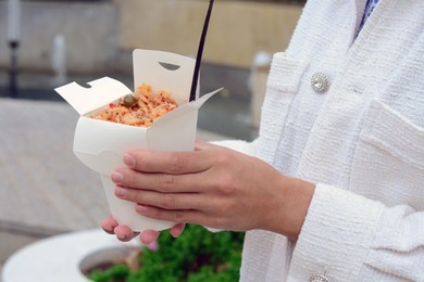 Woman holding paper box of takeaway noodles with fork outdoors, closeup. Street food
