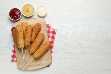 Delicious deep fried corn dogs with sauces on white wooden table, flat lay. Space for text