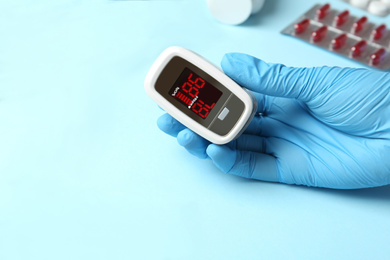 Photo of Doctor in latex gloves holding fingertip pulse oximeter near stethoscope and pills on light blue background, closeup