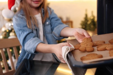 Photo of Little child in Santa hat taking baking sheet with Christmas cookies out of oven indoors, closeup