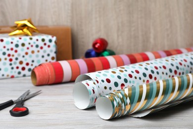 Different colorful wrapping paper rolls and scissors, on white wooden table, closeup