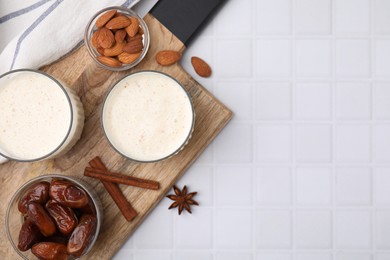 Photo of Glasses of delicious date smoothie, dried fruits, spices and nuts on white tiled table, flat lay. Space for text