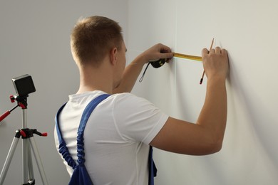Worker using cross line laser level, pencil and tape for accurate measurement on light wall, back view
