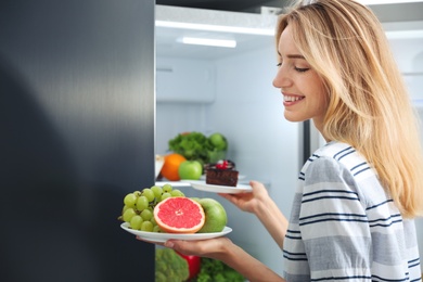 Photo of Choice concept. Woman taking plates with fruits and cake from refrigerator in kitchen