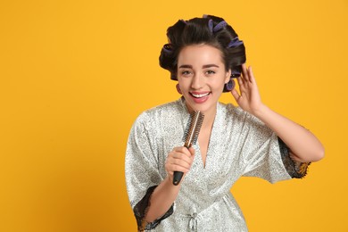 Happy young woman in silk bathrobe with hair curlers singing into hairbrush on orange background, space for text