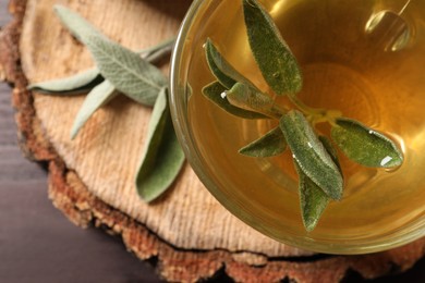 Cup of aromatic sage tea with fresh leaves on wooden stump, closeup