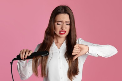 Upset young woman with flattening iron showing thumb down on light pink background. Hair damage