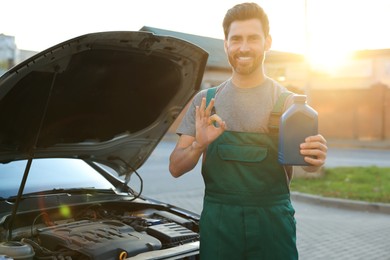 Photo of Smiling worker holding blue container of motor oil and showing OK gesture near car outdoors