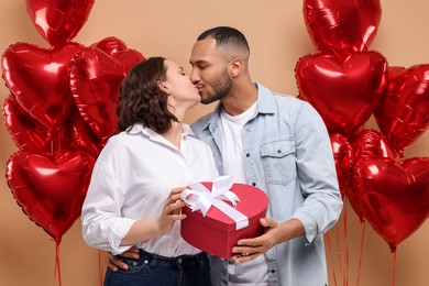 Photo of Lovely couple with gift box near heart shaped air balloons kissing on beige background. Valentine's day celebration