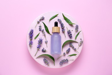 Bottle of lavender essential oil surrounded by leaves and flowers on pink background, top view