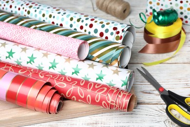 Different colorful wrapping paper rolls, scissors and ribbons on white wooden table