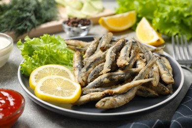 Photo of Plate with delicious fried anchovies, lemon, lettuce leaves and sauces served on light grey table, closeup