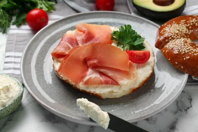 Delicious bagel with cream cheese, jamon, tomato and parsley on white marble table, closeup