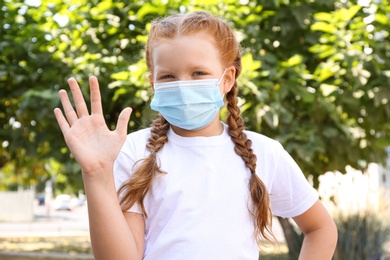 Photo of Little girl in protective mask showing hello gesture outdoors. Keeping social distance during coronavirus pandemic