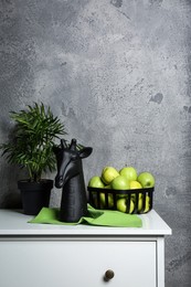 Stylish decor, basket with apples and houseplant on chest of drawers near grey wall indoors, space for text. Interior design
