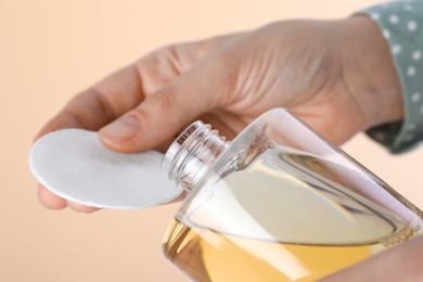 Woman pouring micellar water from bottle on cotton pad against beige background, closeup