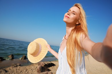 Beautiful young woman with straw hat taking selfie near sea on sunny day in summer